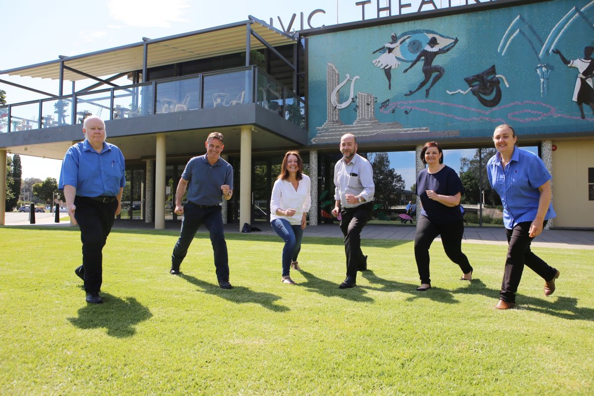 3 men and 3 women taking a step forward on grass in front of Wagga Civic Theatre