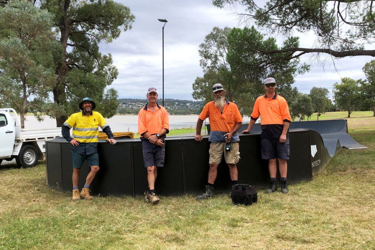 Four men in hi-vis vests standing beside mobile modular pump track with lake in background