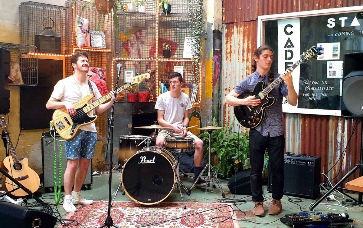 Three young men in courtyard, one with bass guitar, one on drum kit and one with guitar