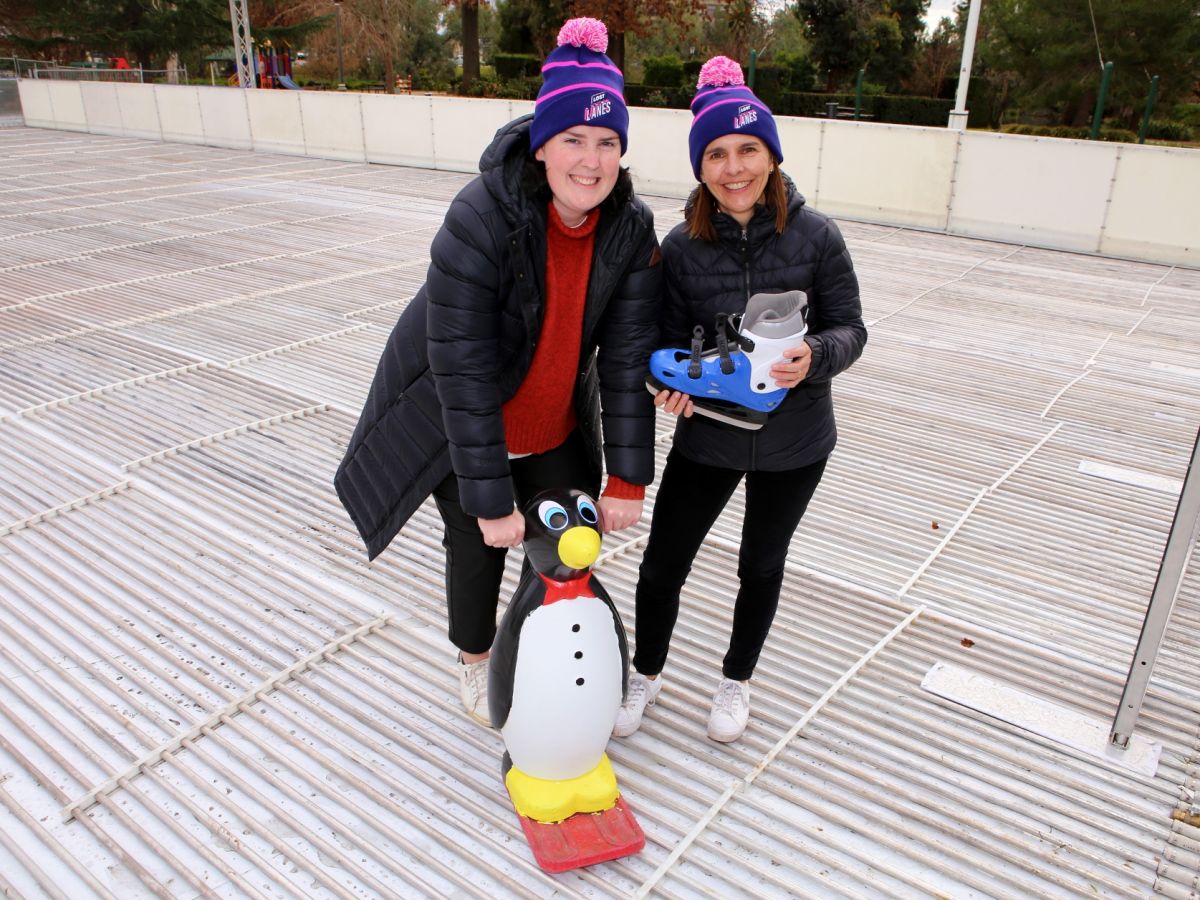 Two women wearing beanies and scarves, holding ice skates, standing on outdoor ice skating rink