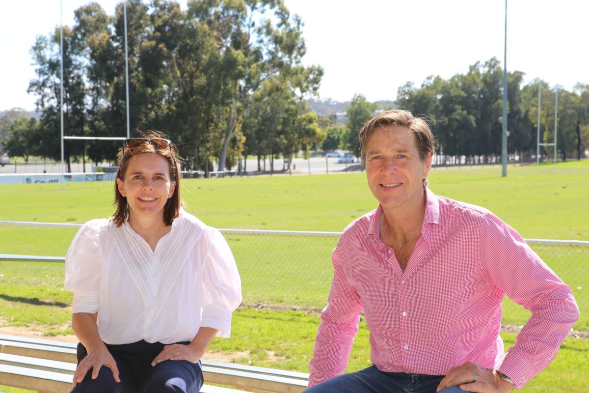 woman and man sitting on sportsground seating with rugby goal posts in background