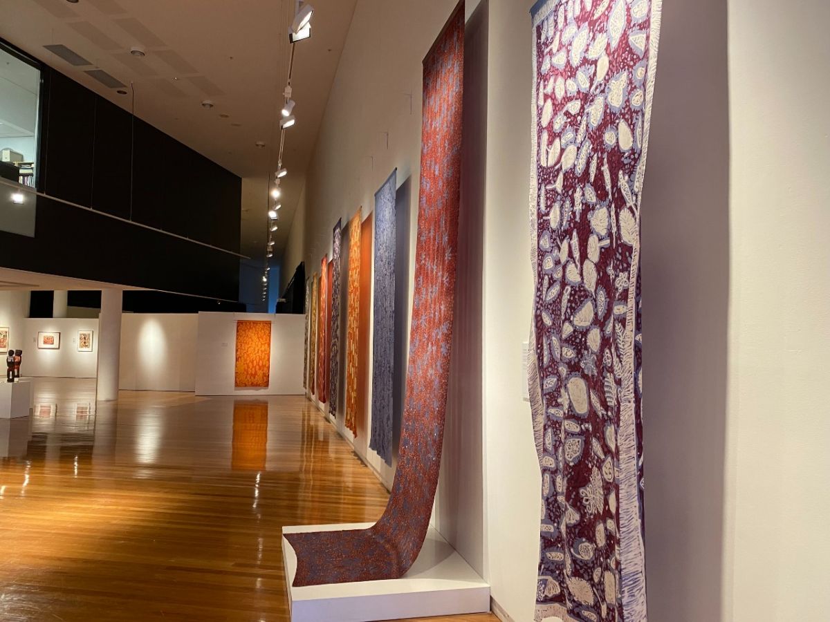 A series of long swathes of decorated fabric hang on a gallery wall