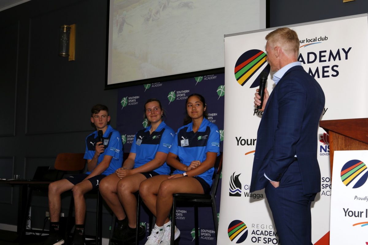 Three teenage athletes in uniform sitting in room, being asked questions by man with microphone