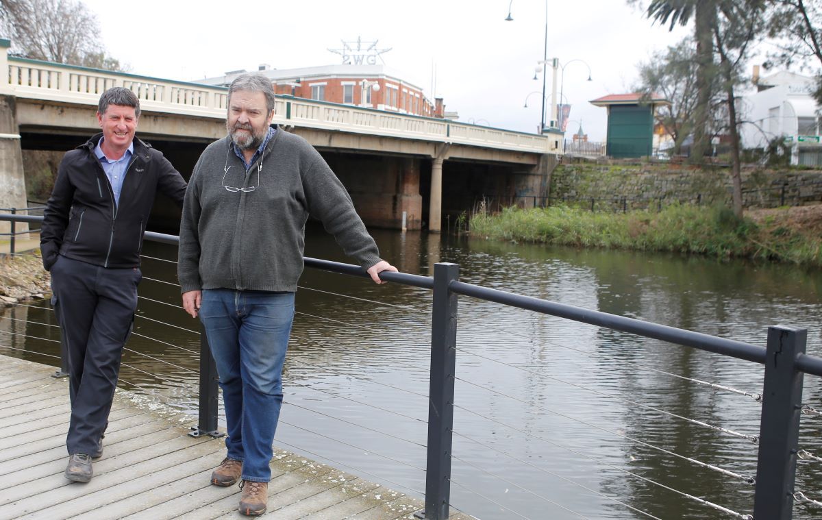 Two men standing on a boardwalk near a body of water. 