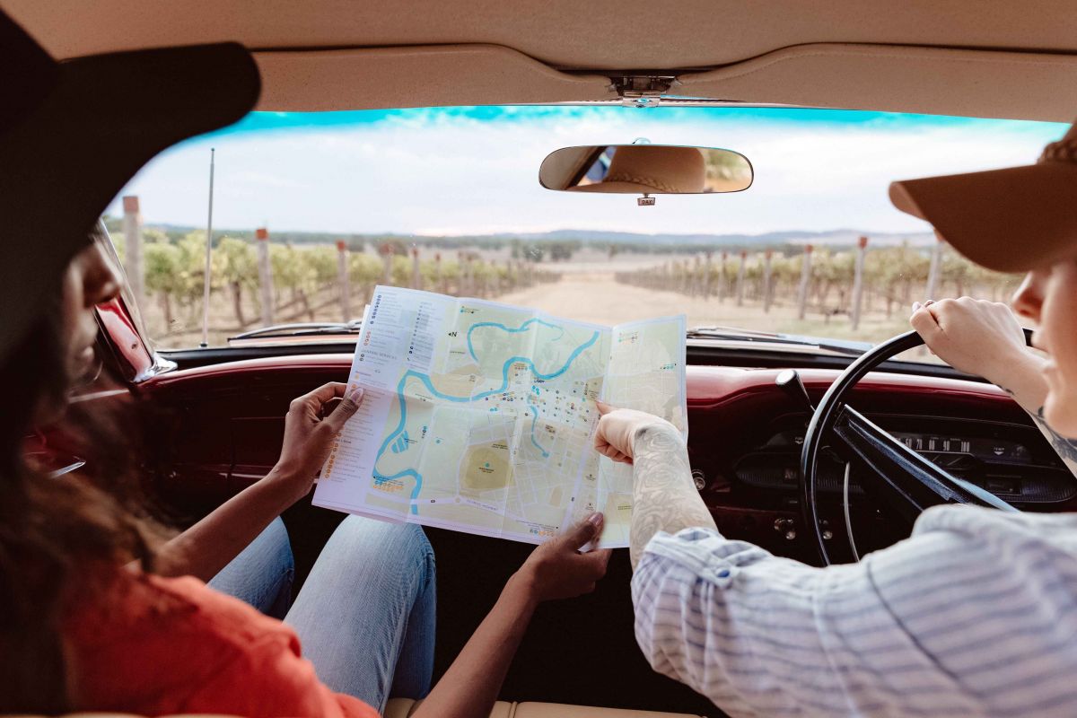 Back view of two people sitting in front seat of car looking at an open map of Wagga Wagga. View out the windscreen of surrounding grape vines at vineyard.