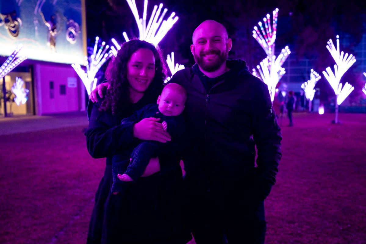A man and woman and an infant standing in front of a field of illuminated trees. 