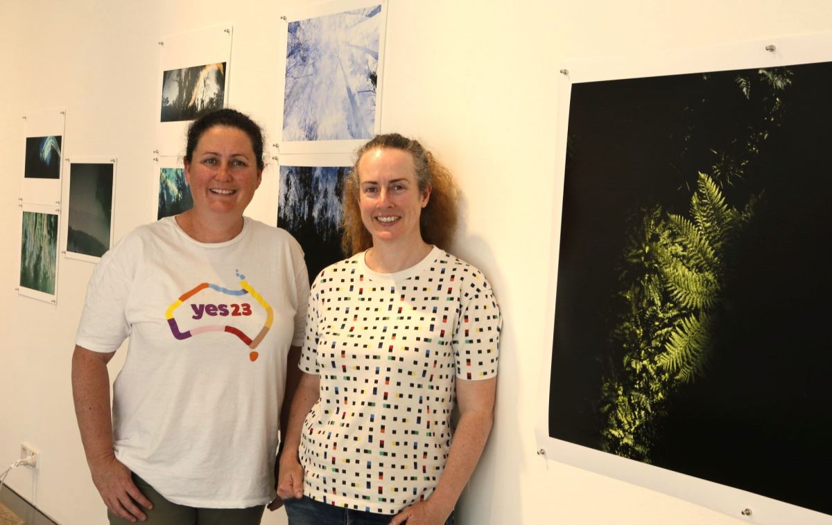 Two women standing with photographs of landscapes which are pinned to a gallery wall. 