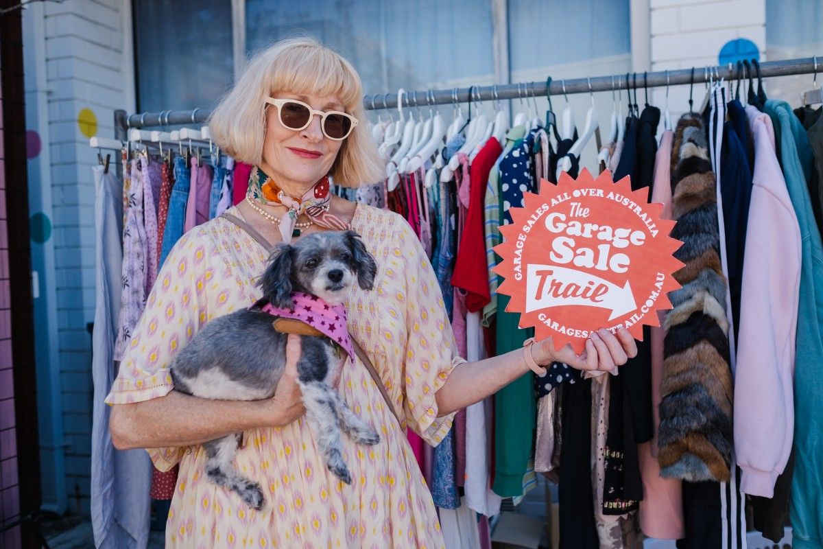 A lady wearing sunglasses holds a small dog under her right arm while holding a sign in the her left hand. The sign reads 'The Garage Sale Trail'. 