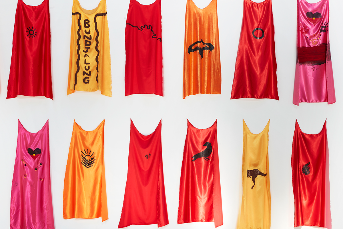 A series of silk capes on display on a wall. They are various warm colours and have hand painted symbols on them.