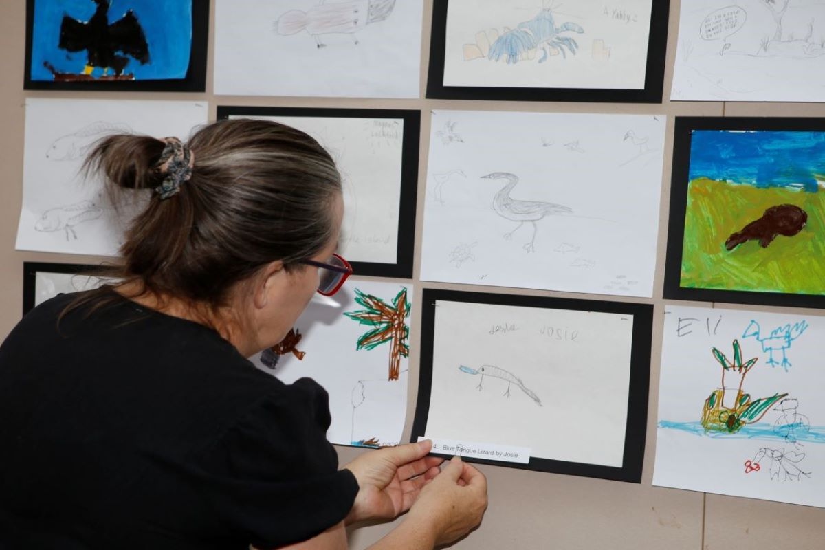 A woman pins a child's drawing onto a wall where there are already other children's artworks. 