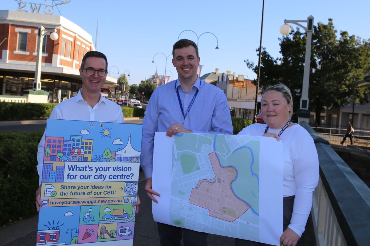 Two men and a woman stand smiling in a street. They are holding and sign and map with information about the CBD Masterplan.