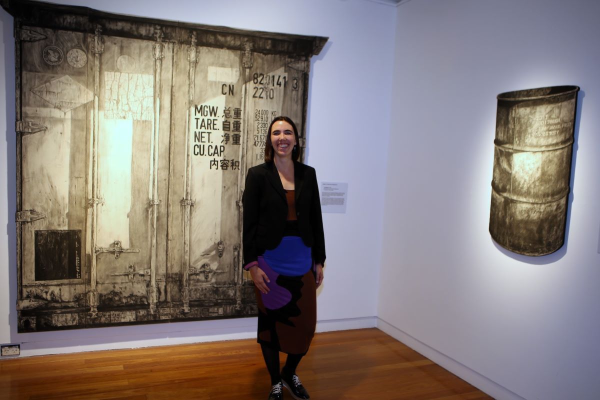 A woman stands in an art gallery space with her artworks that are black and white large scale drawings. 