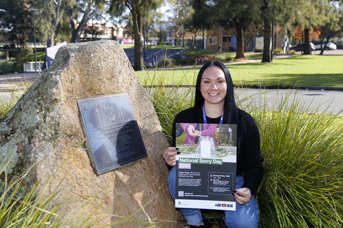 Woman holding a poster about National Sorry Day, while crouched next to the Sorry Day Rock in Wagga Wagga.