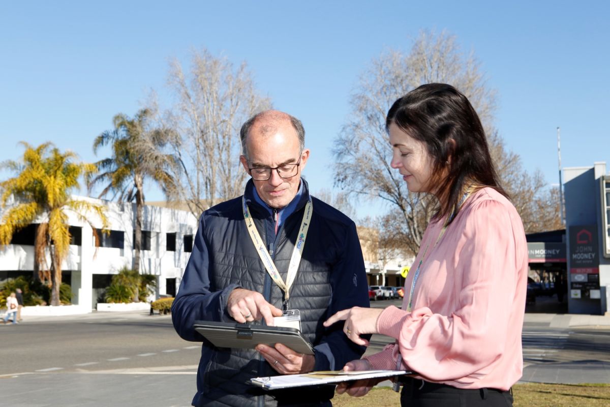 A man is holding an Ipad and a woman is holding a printed survey and clipboard. They are both outside.