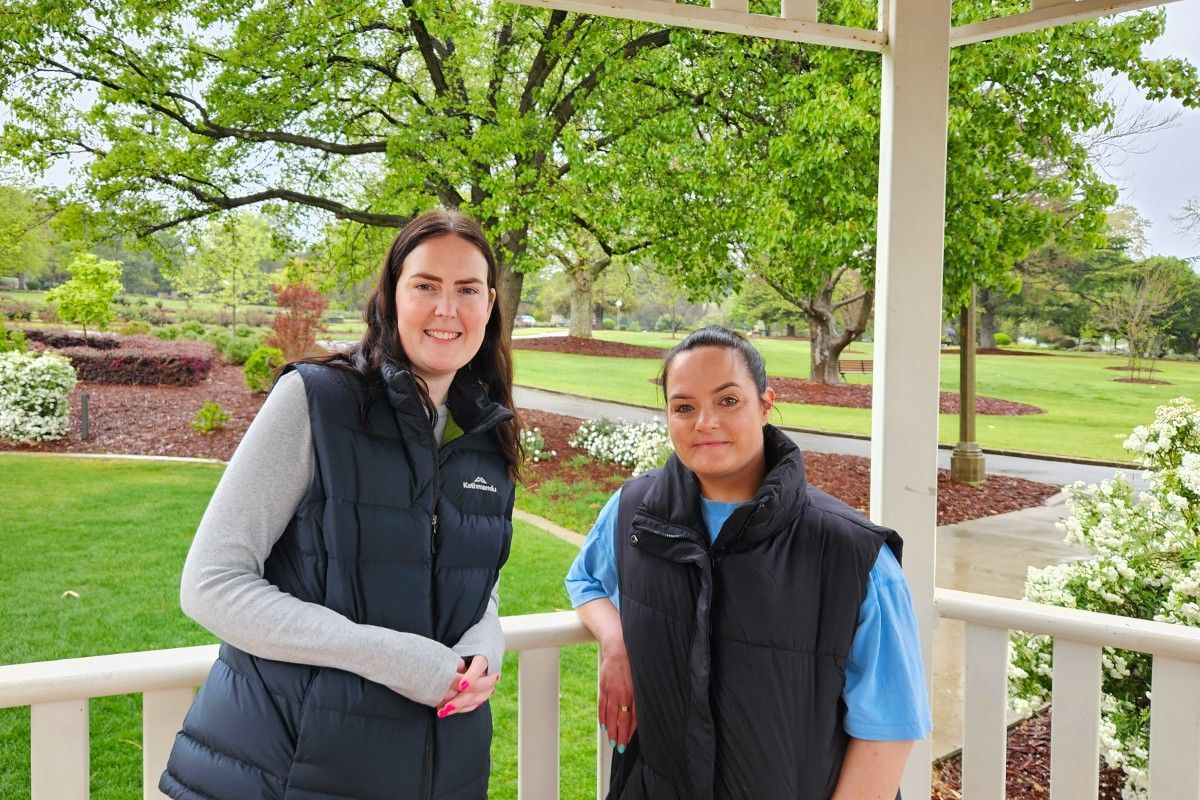 Two women wearing vests lean against the railing on a rotunda, with public gardens in the background.