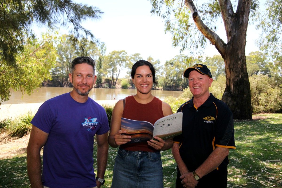 Two men and a woman standing near the banks of a river, holding a tourism guide