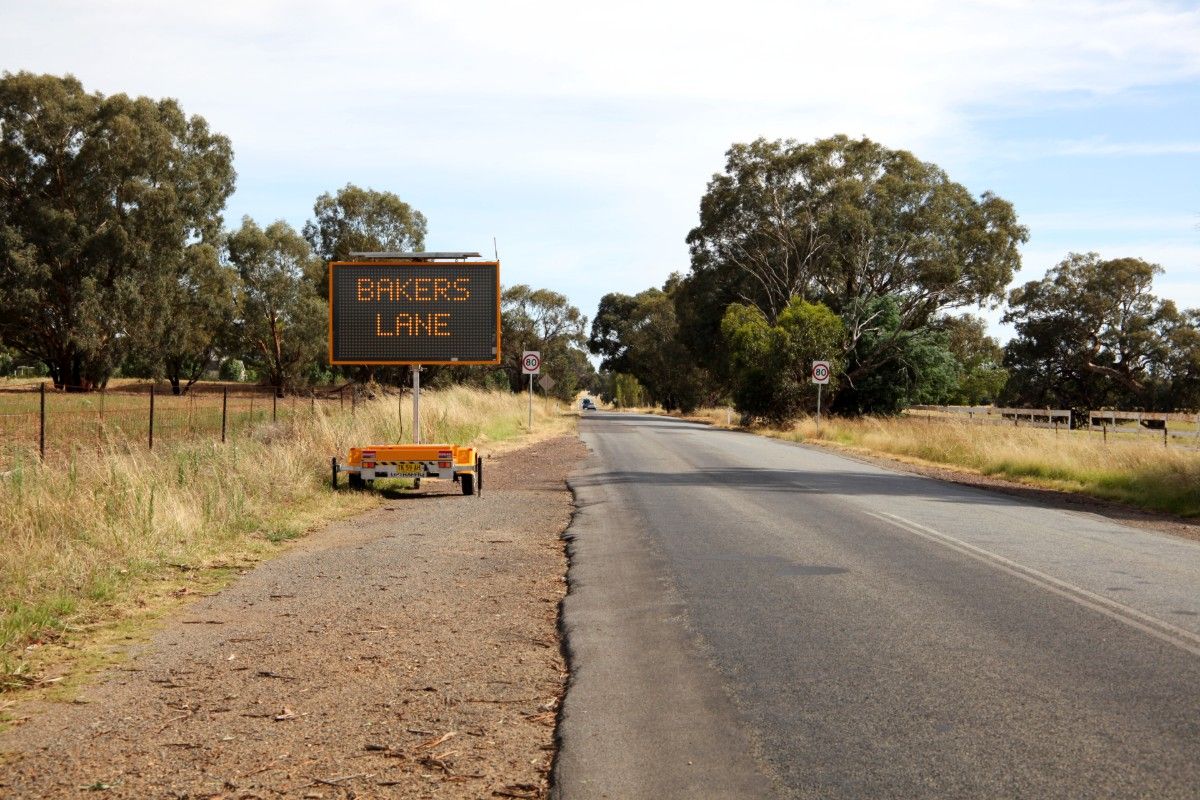 Roadside electronic messaging board saying Bakers Lane next to a bitumen road, with 80-k signs and traffic in the background.