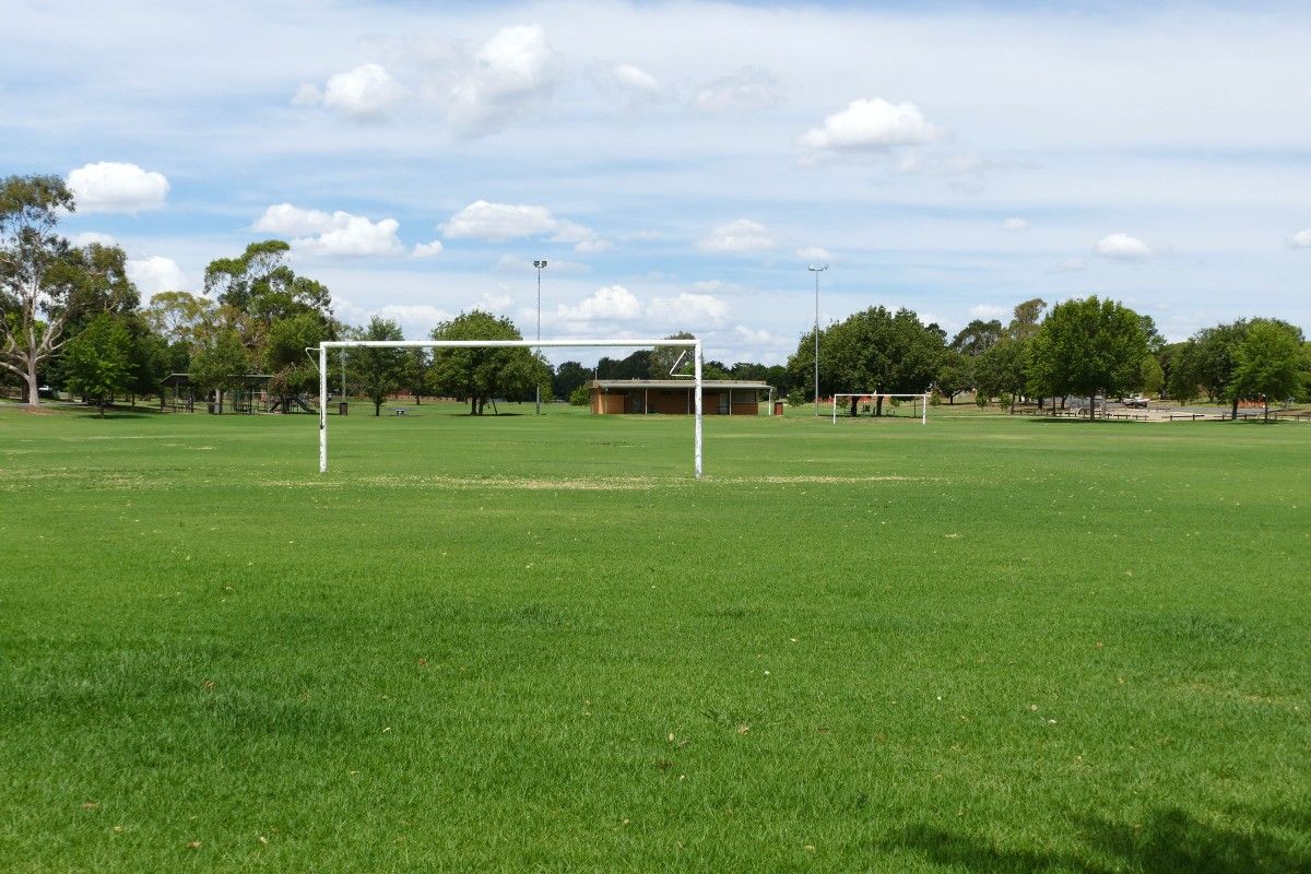 Soccer goal post frame in foreground, with green grass sporting oval and brick amenities building in the background.