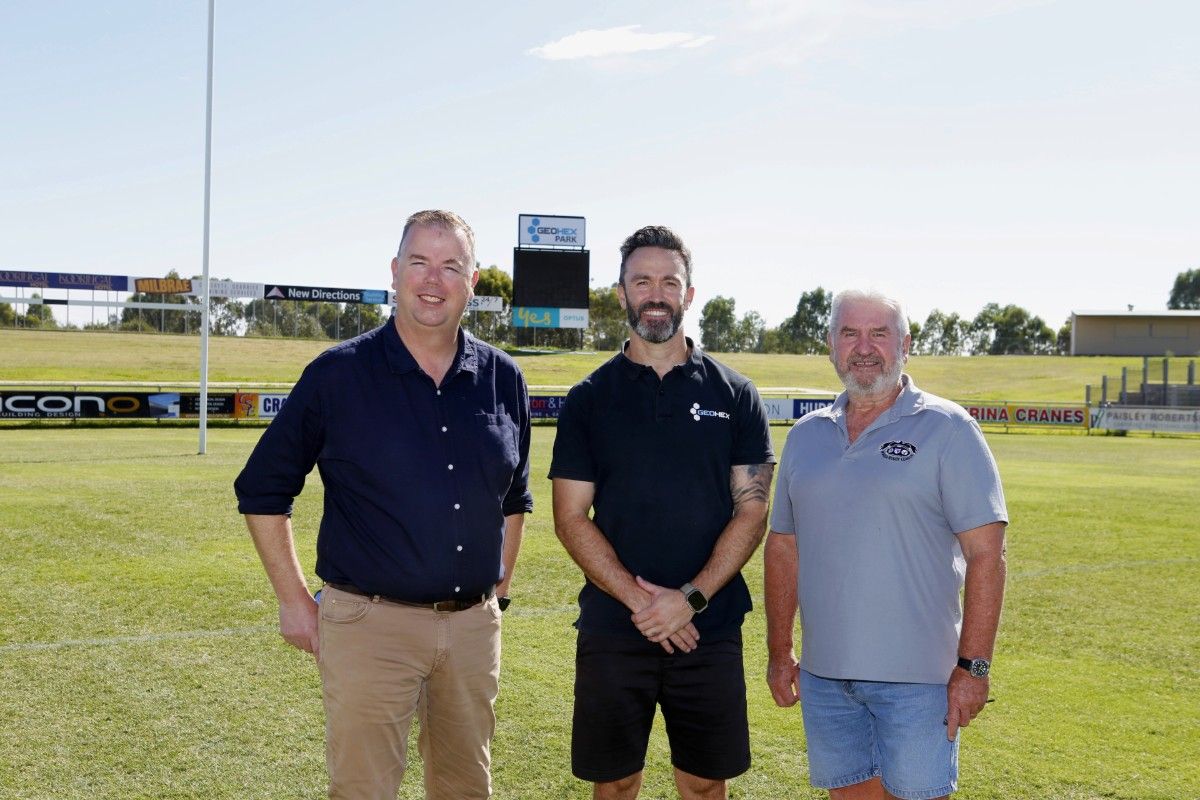 Three men standing on a rugby league field, with goal posts and a scoreboard in the background. 