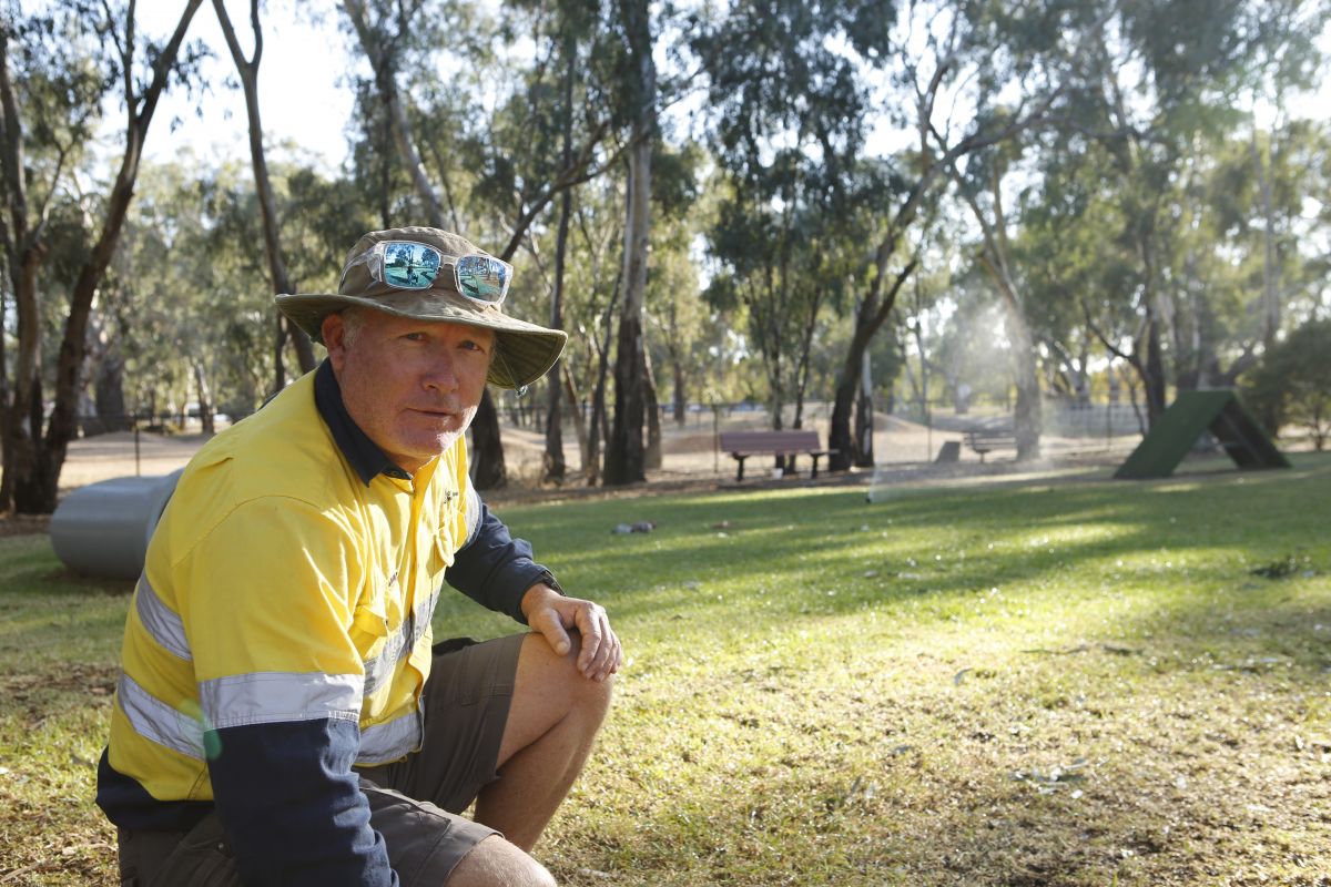 Man wearing a hi-vis shirt, hat and sunglasses crouches on turfed lawn area of a dog park, with sprinklers watering the turf in the background.
