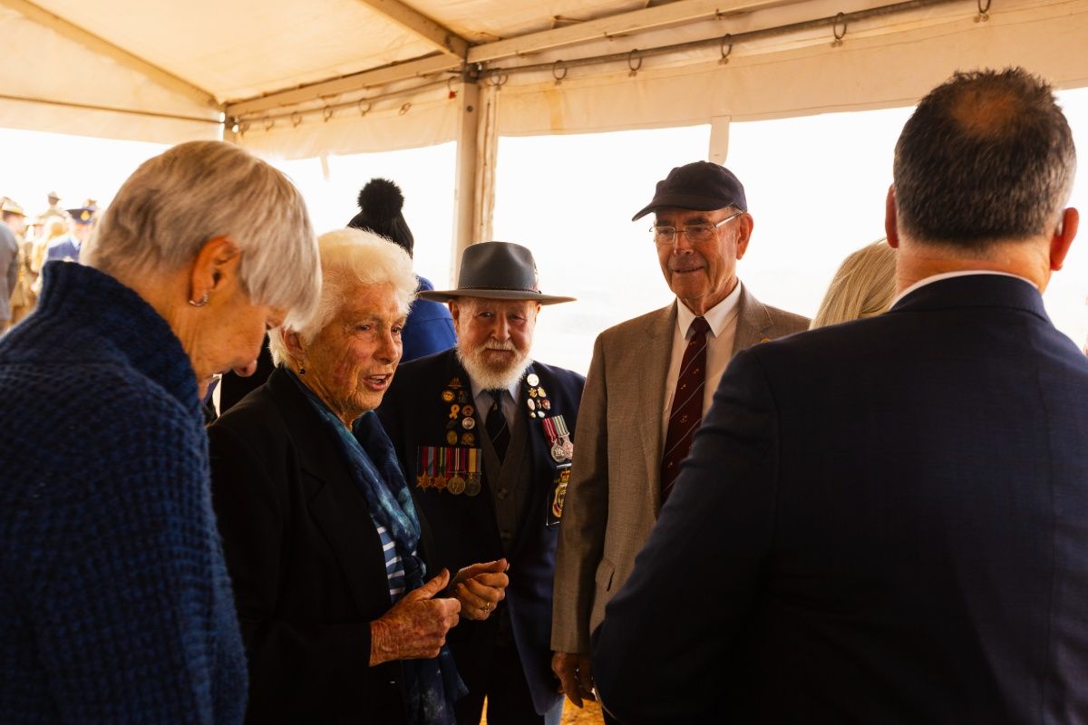 : An elderly woman named Patricia Schlitz, stands dressed in navy blue and in conversation with another woman and three men, smiling as she talks with them. One of the men wears a broad brimmed hat and has multiple brightly coloured ribbons and military service medals pinned to the lapels of his navy coat. The man to his right wears a black cap, reading glasses and a light-coloured suit with a maroon tie. 