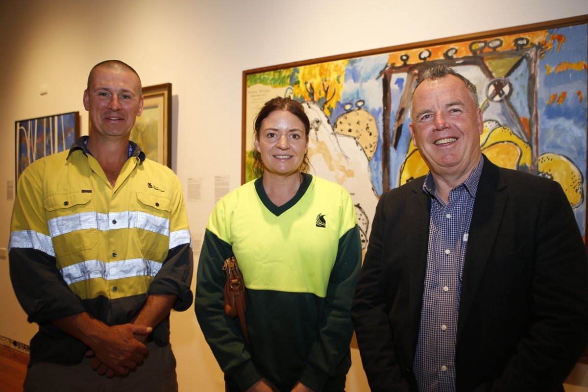 A man and woman in high vis work shirts with a man in a suit, all standing in front of an artwork handing on the wall of an art gallery.