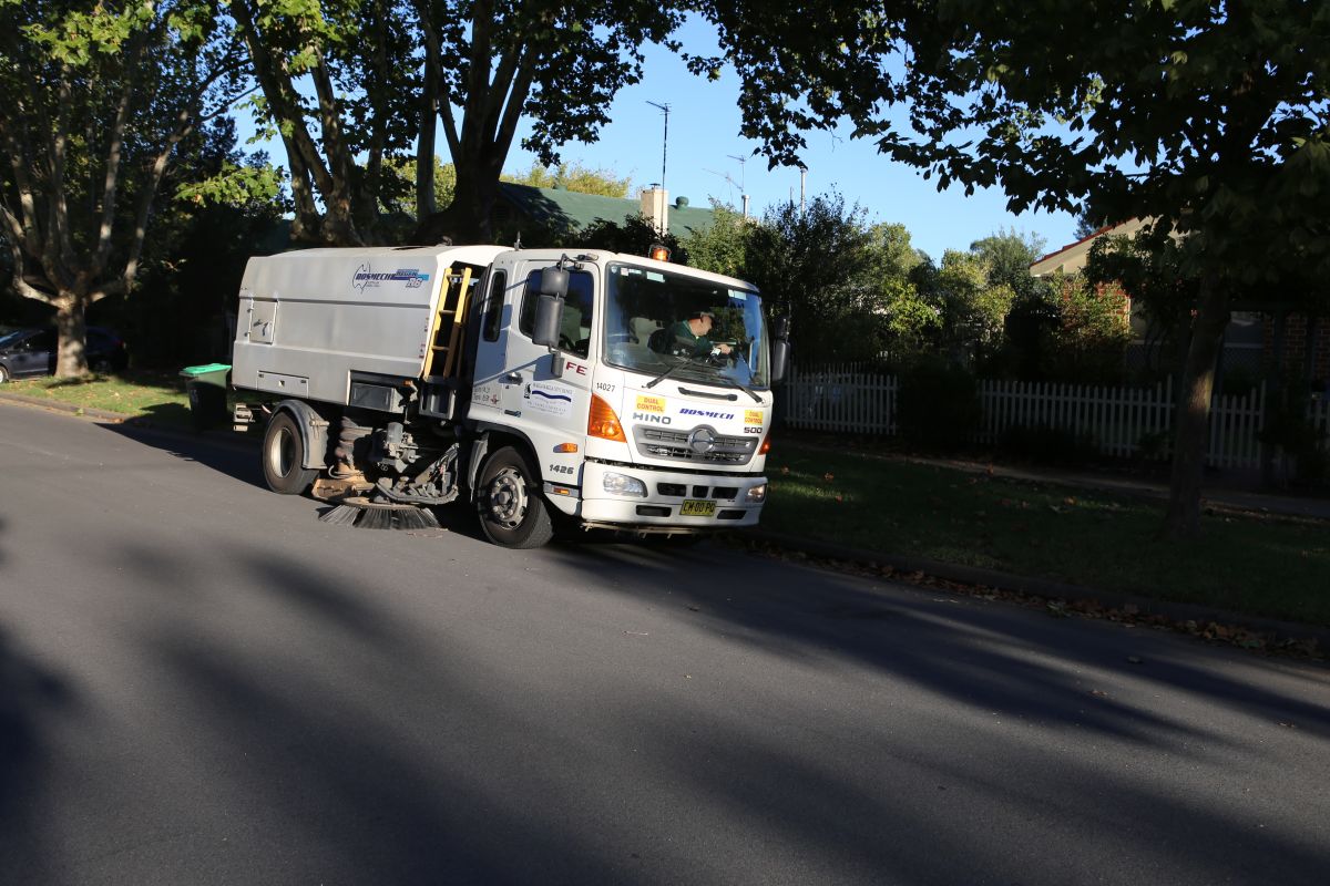 A white street sweeper vehicle situated on the roadside, of a suburban street lined with leafy trees.