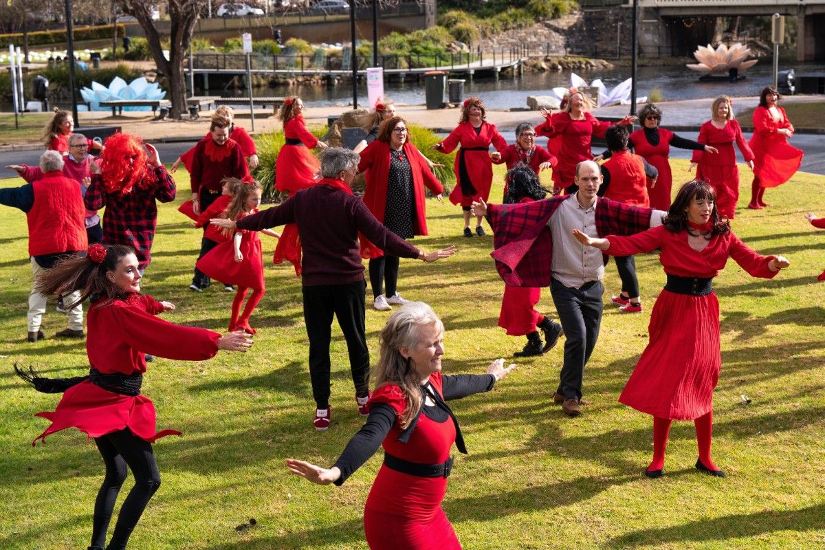 A group of men, women and children all dressed in red, dancing on a grass lawn area.