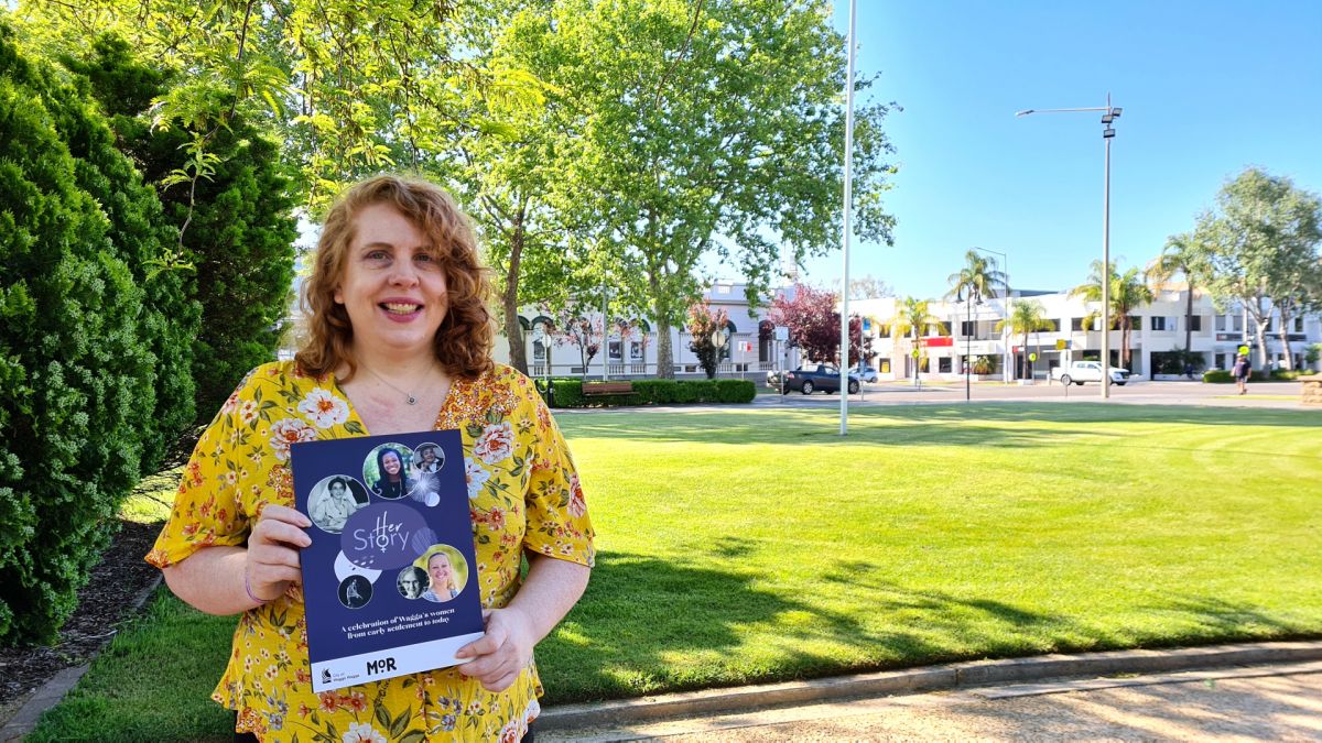 Woman holding book, HerStory, standing park with Museum of Riverina and Wagga Civic Centre in background