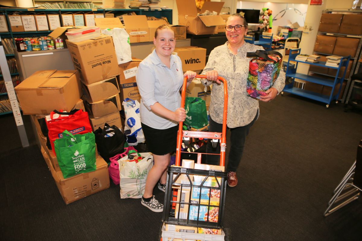 two women standing next trolley packed with bags of food items, with boxes of food item next to them