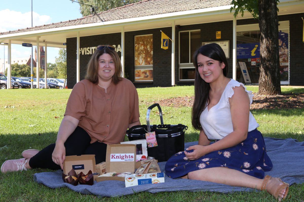 Two women sitting on picnic blanket with basket and food items displayed on blanket, building in background