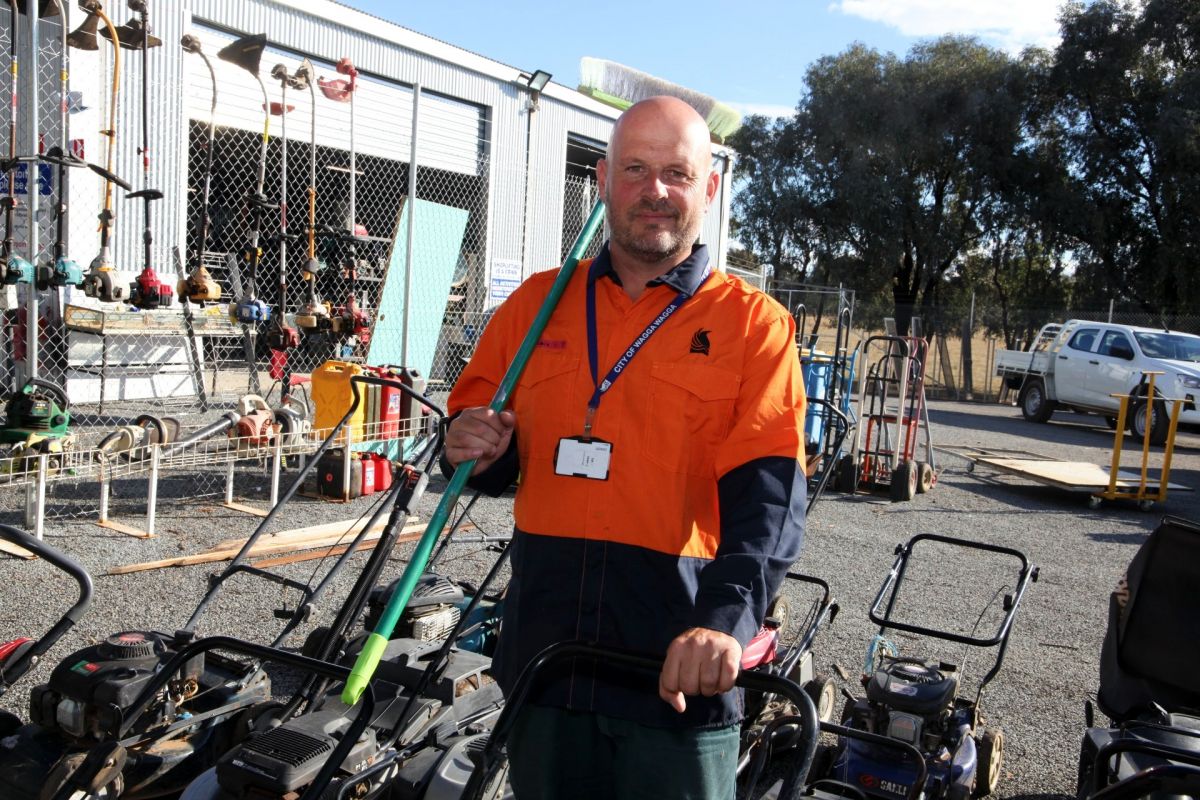 Man in orange flouro vest holding broom, standing next to lawn mowers with whipper snippers in background