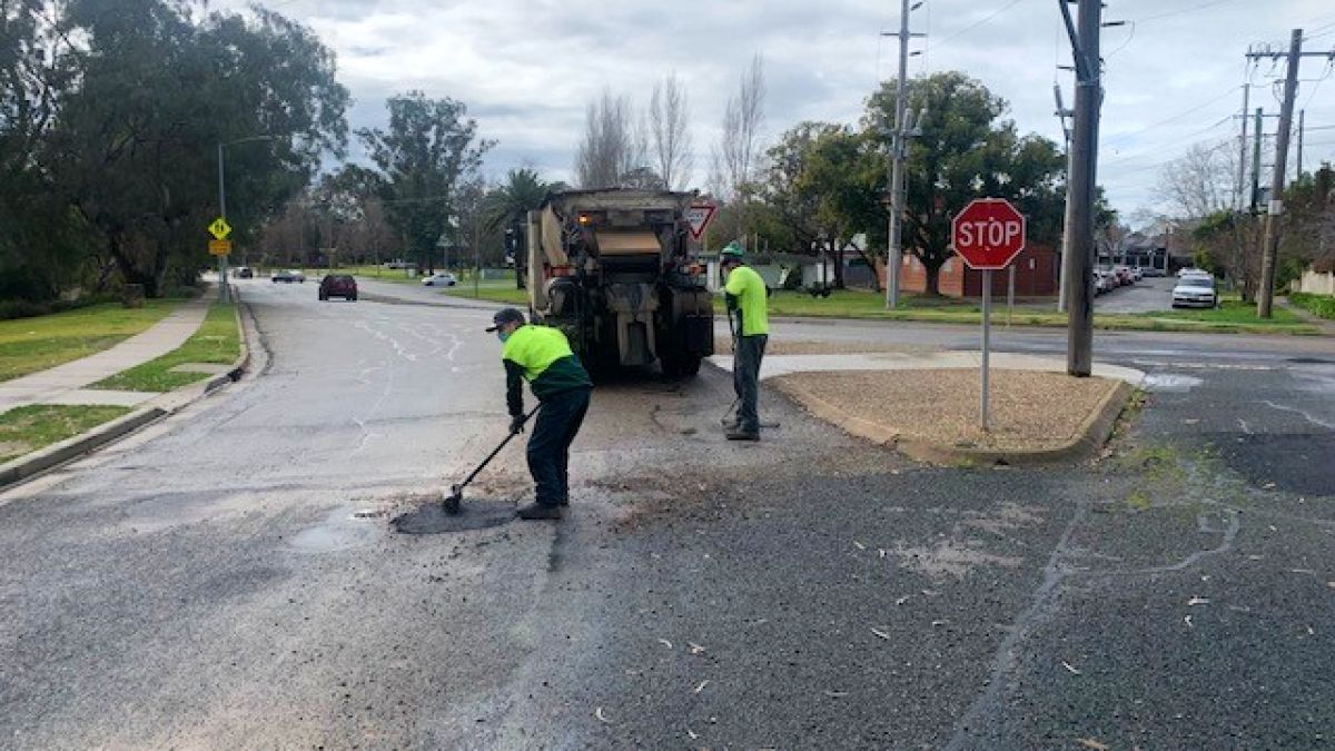 Two men in safety shirts behind truck, filling in potholes