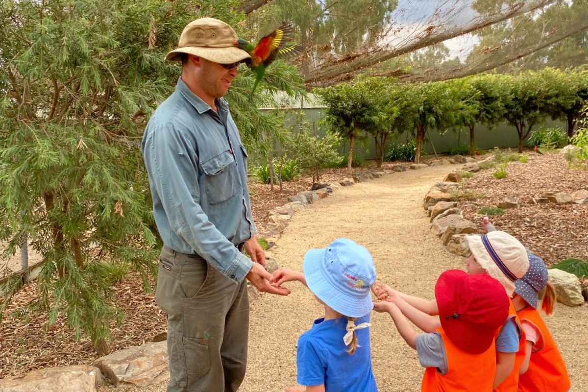 Man with bird on hat with three young kids handing him treats for bird