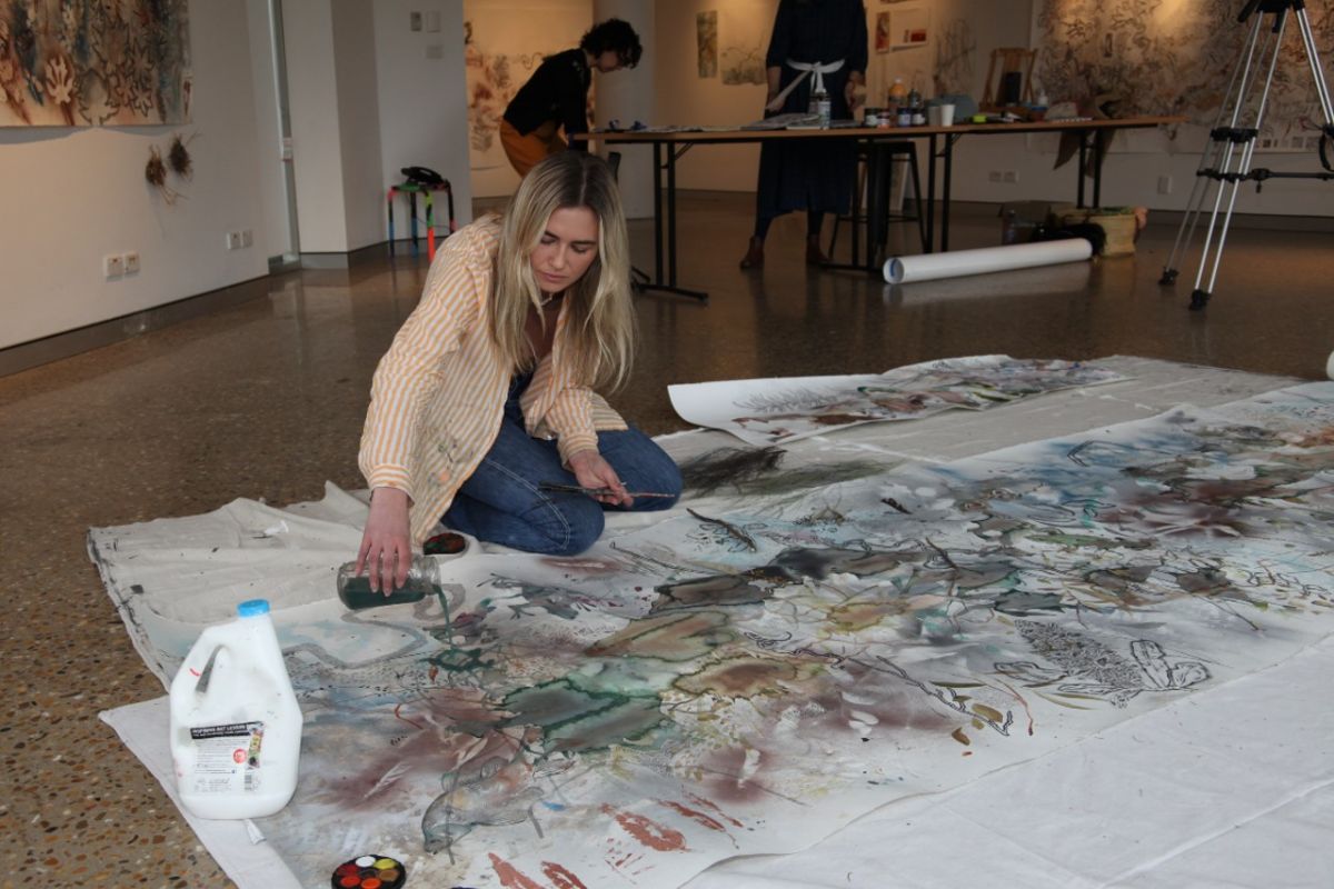 A young woman pours paint onto an artwork laying on the floor