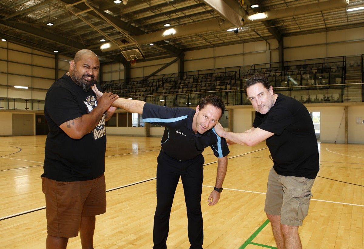 Male wrestler pretending to twist another man's arm, with a third man watching on 