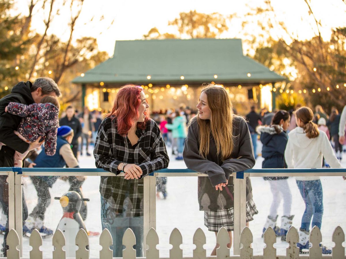 Two women leaning on fence with pop-up ice skating rink and skaters in the background