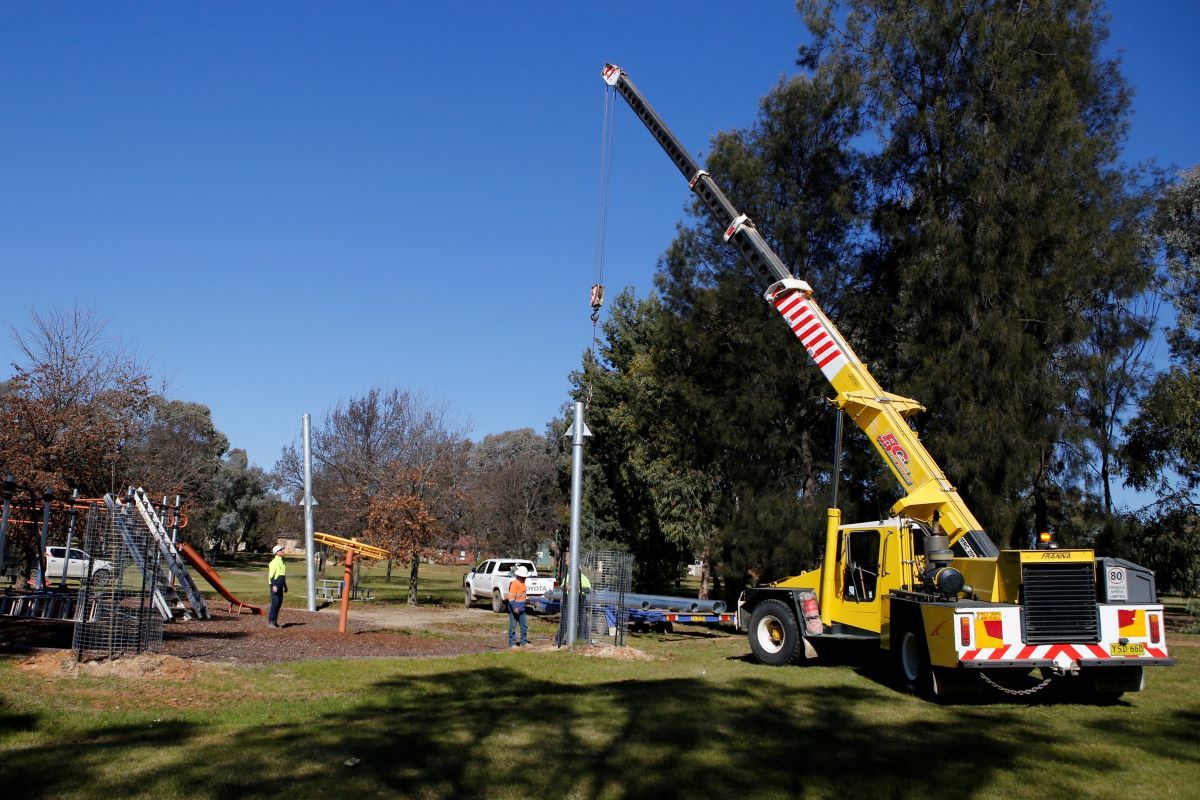 Two men position a large pole, that is being held up by a crane, into the ground next to a playground. 