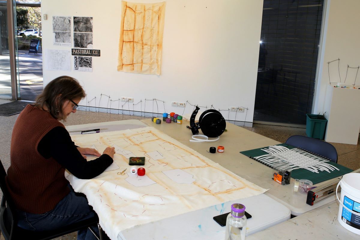 a woman sits at a table in an art space making an artwork out of a large piece of light coloured fabric. 