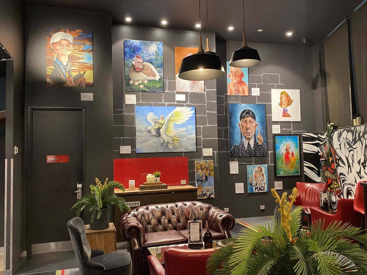 Satirical artworks featuring notable Australians hanging on wall inside gallery space.