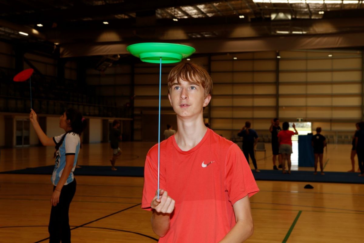 In the foreground a young male student balances a disk atop a thin stick, focusing intently. In the background other students can be same practicing the same skill.