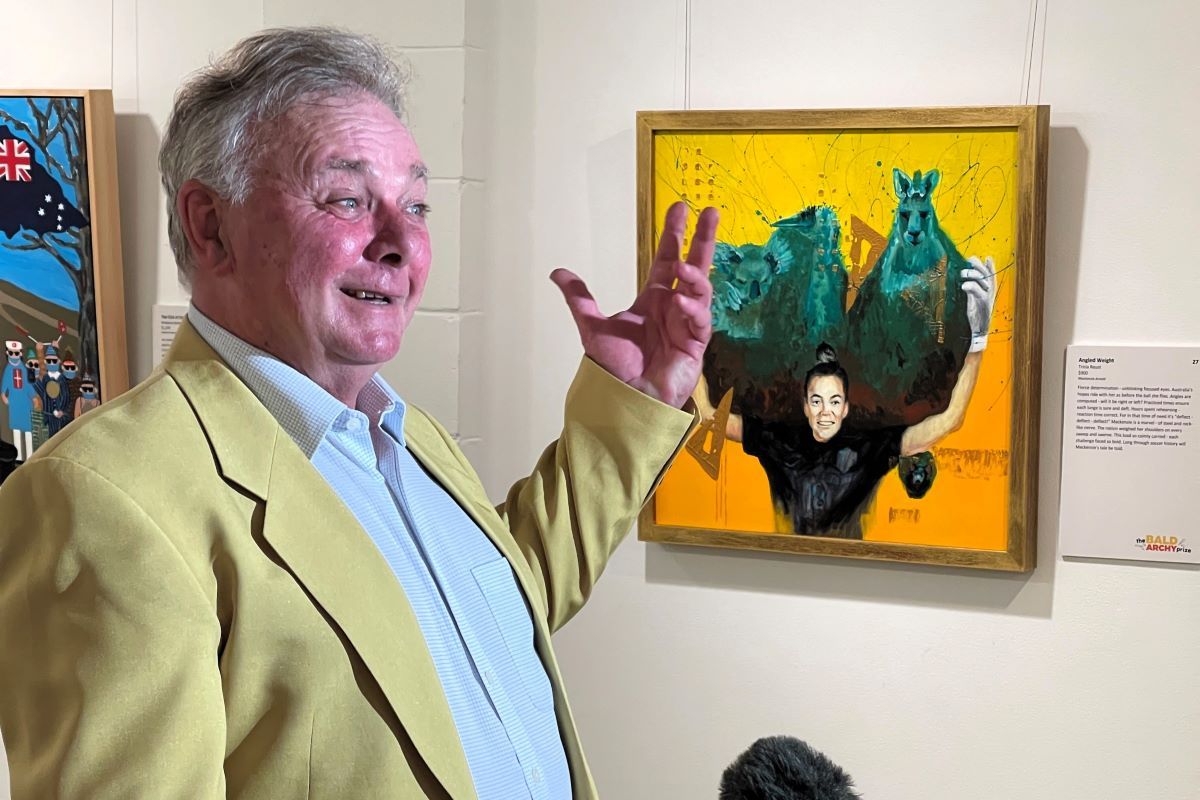 A man stands in front of a portrait in an art gallery. He looks happy and is gesturing with his arm. 