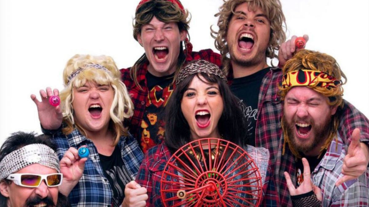 A group of six people are huddled together around a red, hand-held bingo machine wheel. They all wear wigs and bandanas, some wear sunglasses. All have their mouths open to indicate cheering or sounds of excitement.