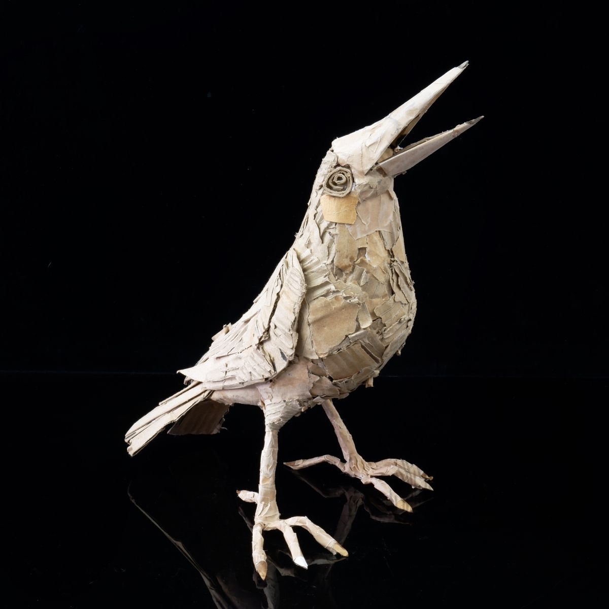 A small bird sculpture standing freely. The bird is made from cardboard and other recycled materials.
