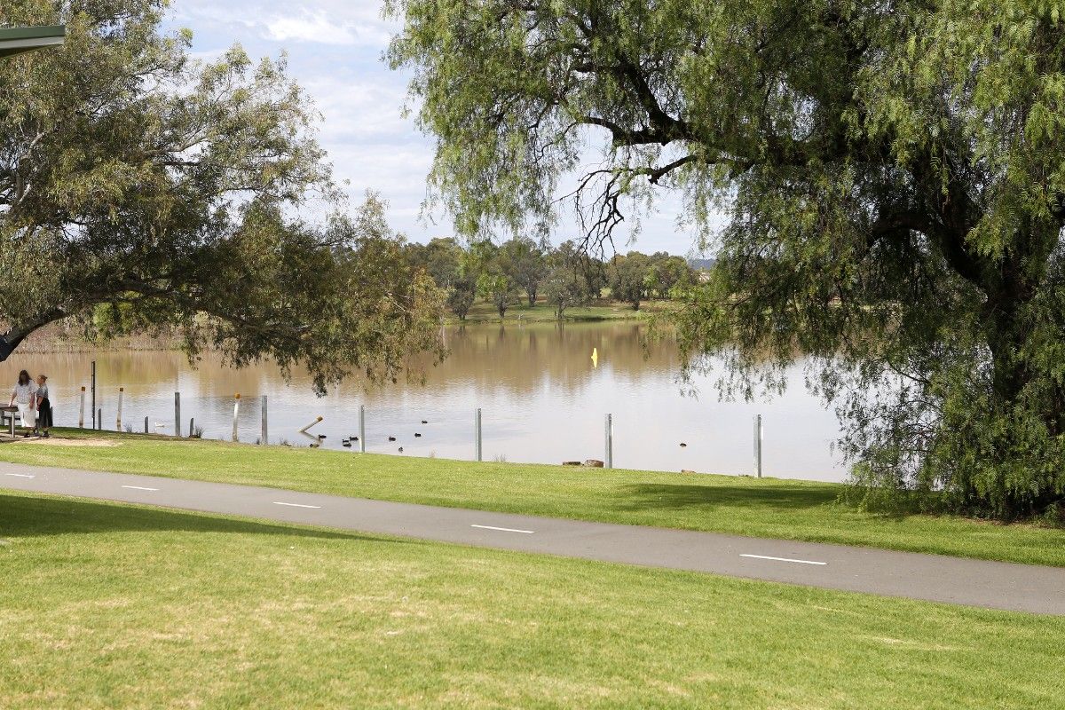 Two women in distance standing next to picnic table beside lake, with grass and bitumen shared pathway in foreground.