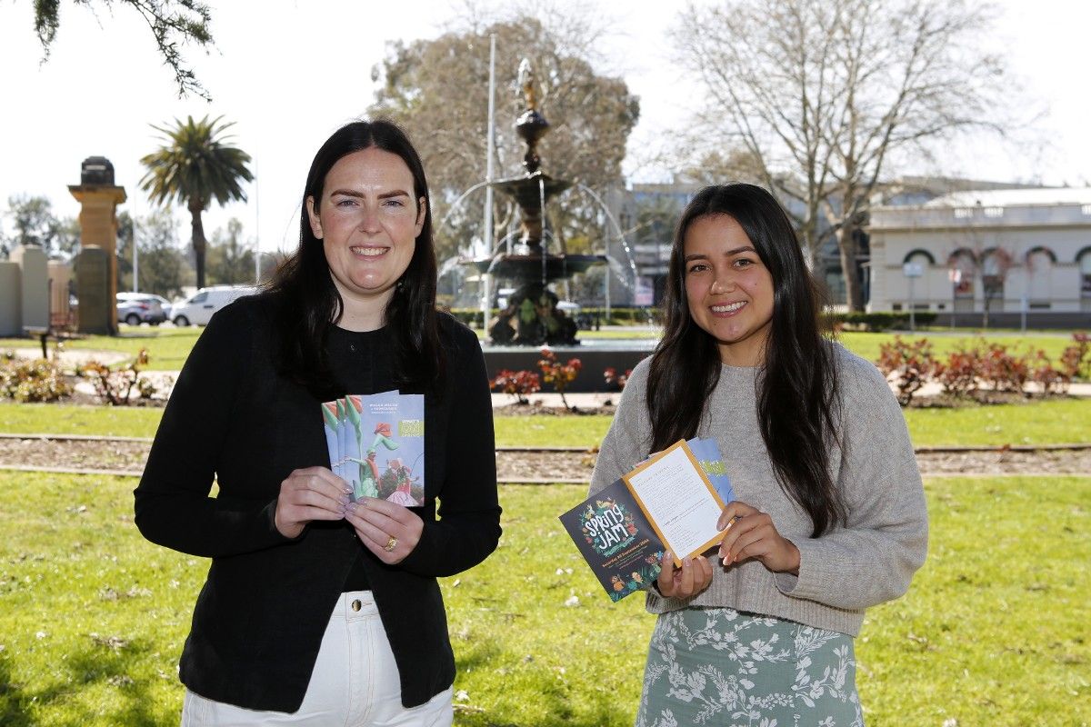 Two women holding small pamphlets with ‘What’s On’ on cover, standing in front of a fountain in a public garden.