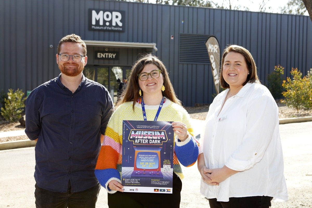 Two women and a man standing outside with main Museum of the Riverina building in the background, and one of the women holding a poster promoting Museum After Dark.