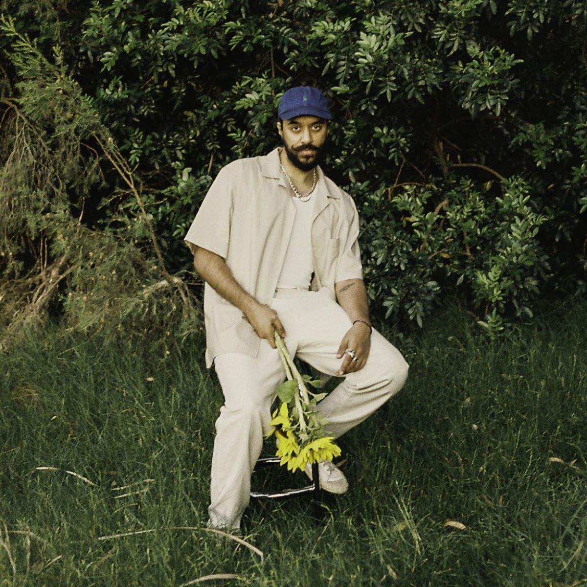 Man with a beard, and wearing a baseball cap and holding flowers, sits on a stall on grass, with trees in the background