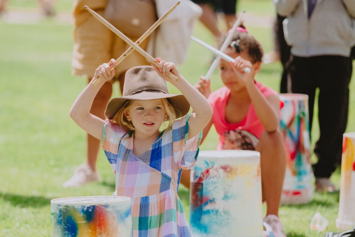 Two young girls holding drum sticks crossed above their heads, with coloured drums on the ground in front of them.