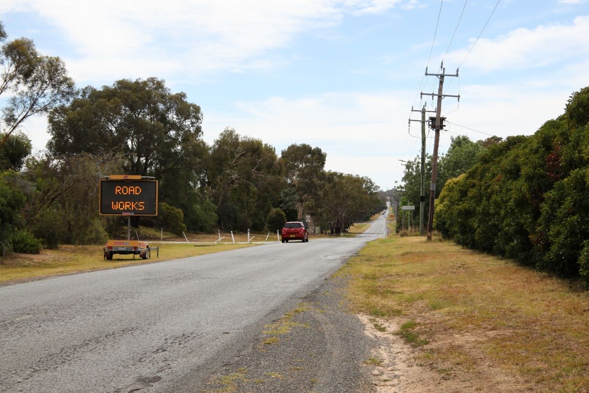 A roadside electronic messaging board stating Road Works next to a road, with a red car in the background travelling along the road.